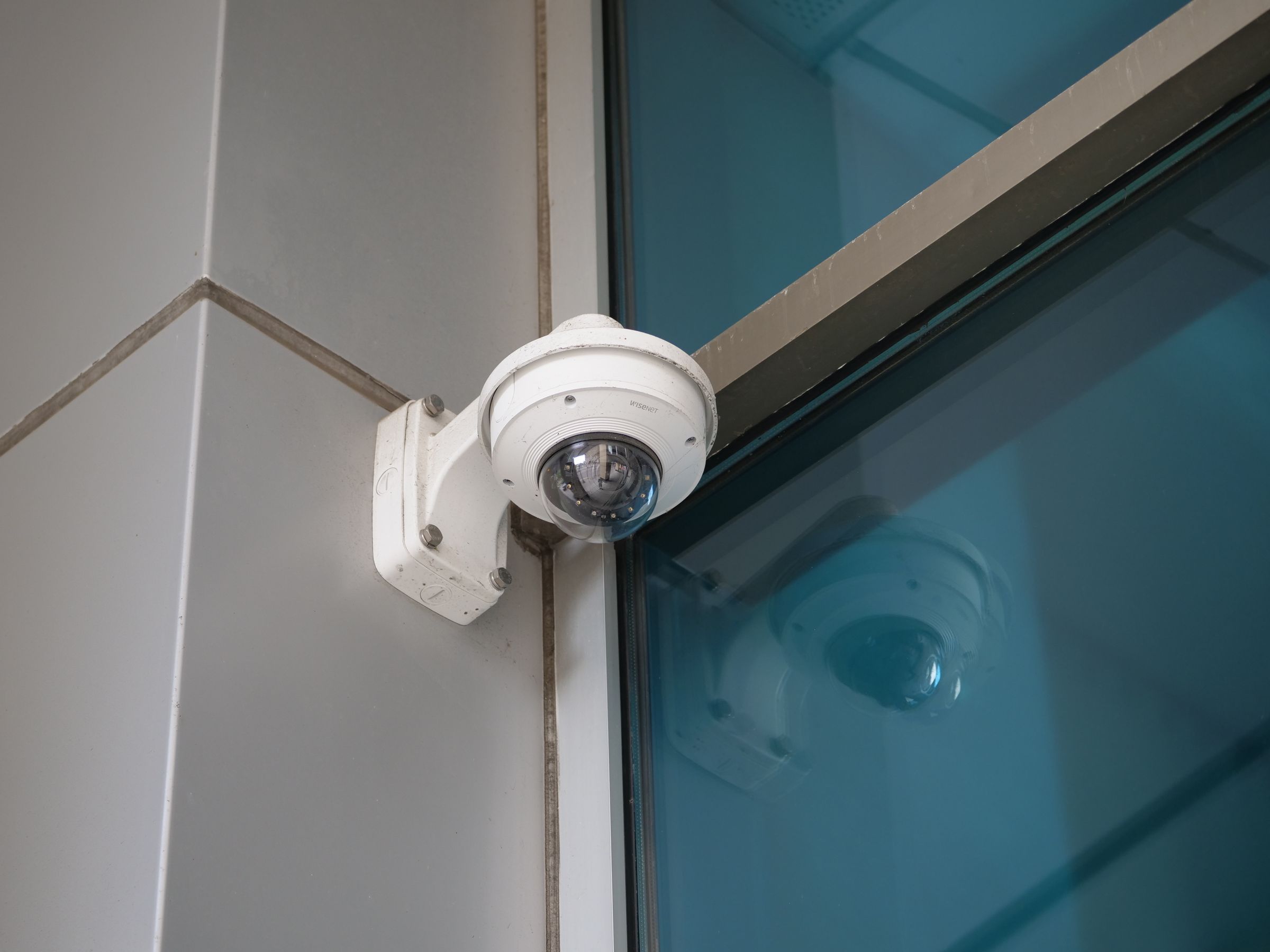 Câmara de videovigilância tipo dome fixada numa parede cinzenta, junto a uma fachada de vidro