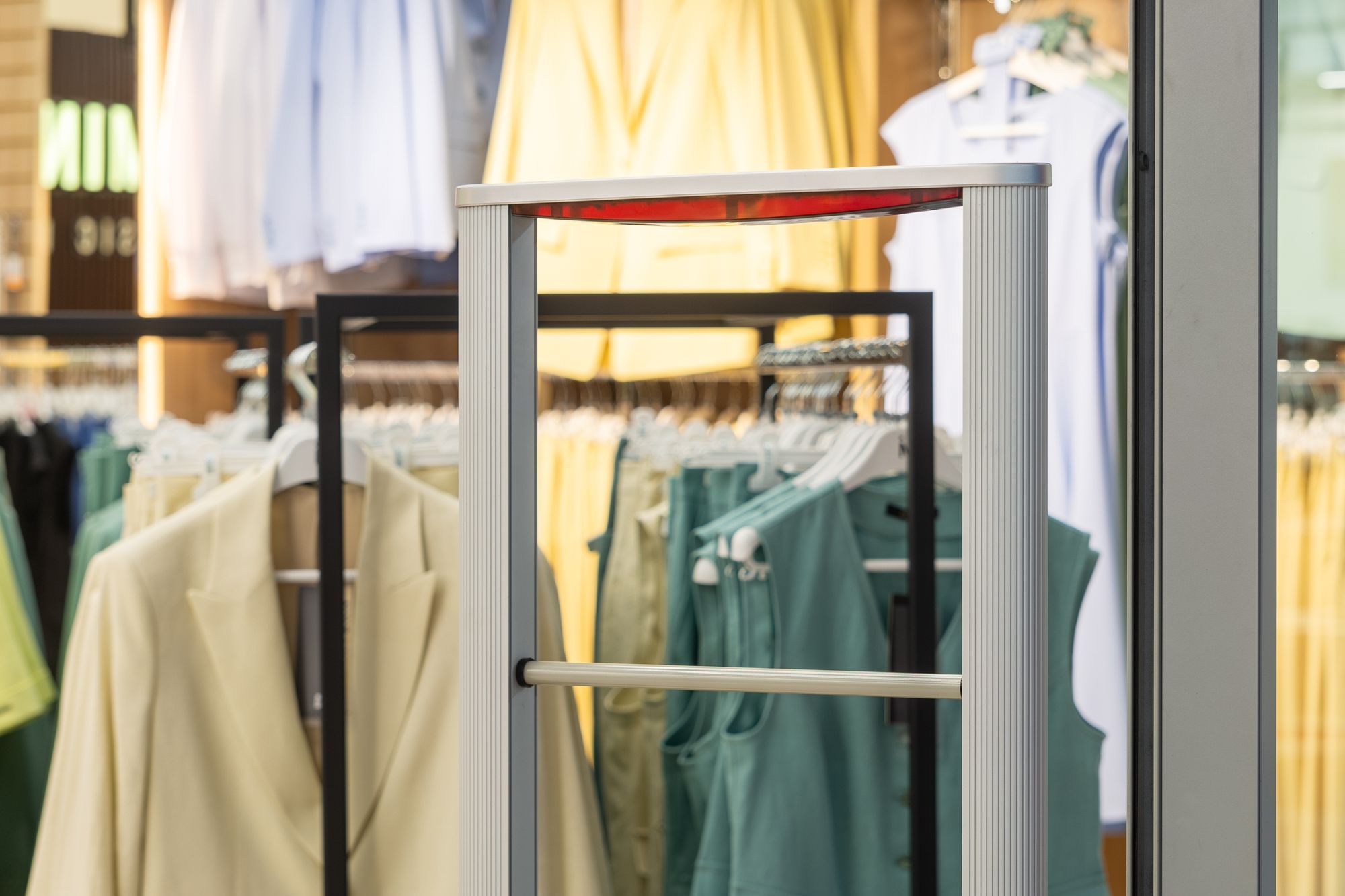 Antena anti-furto com luz vermelha à entrada de uma loja de roupa, com peças em cabides ao fundo