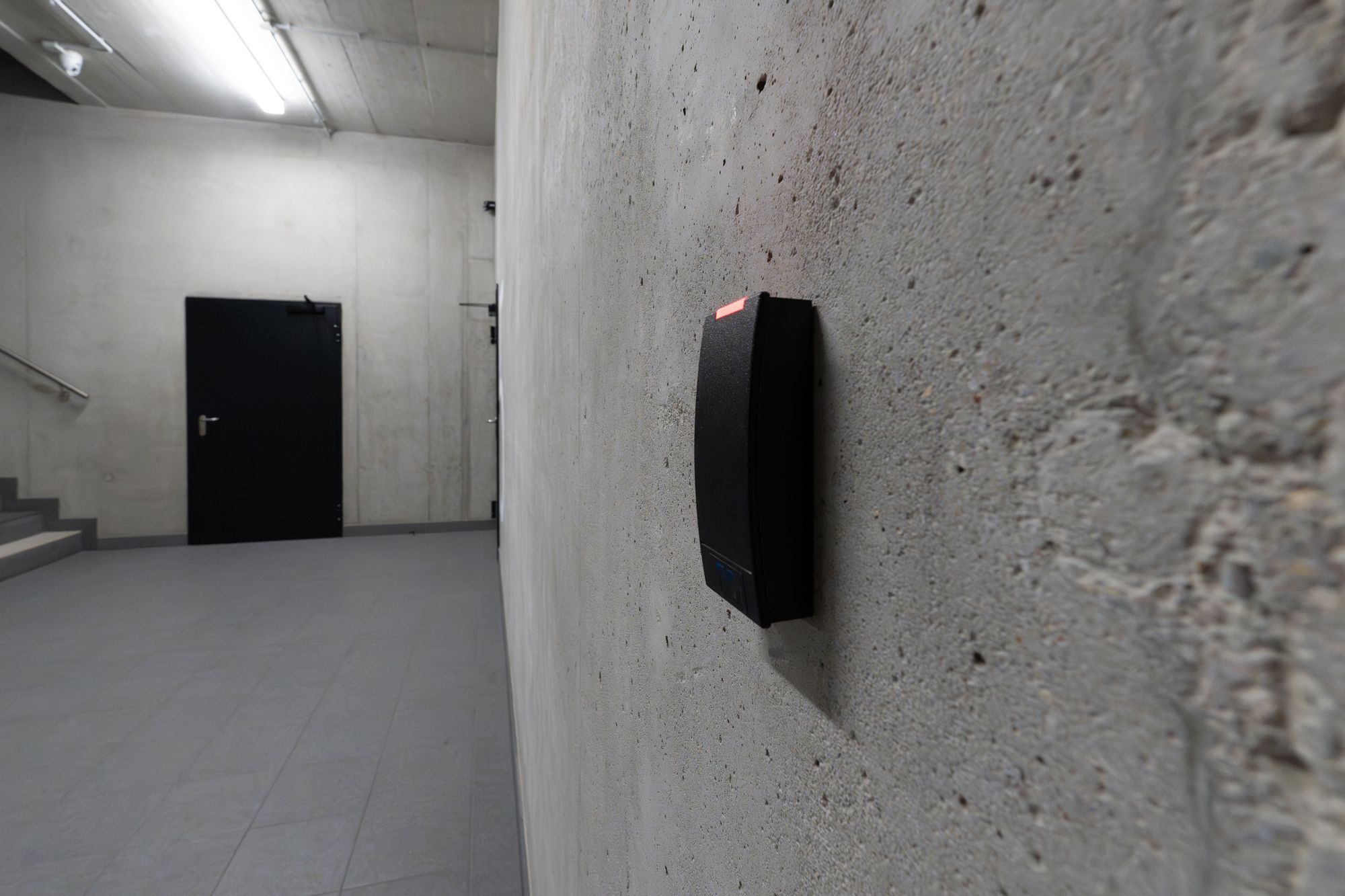 Leitor de cartões preto com LED vermelho fixado numa parede de betão, com porta preta ao fundo do corredor
