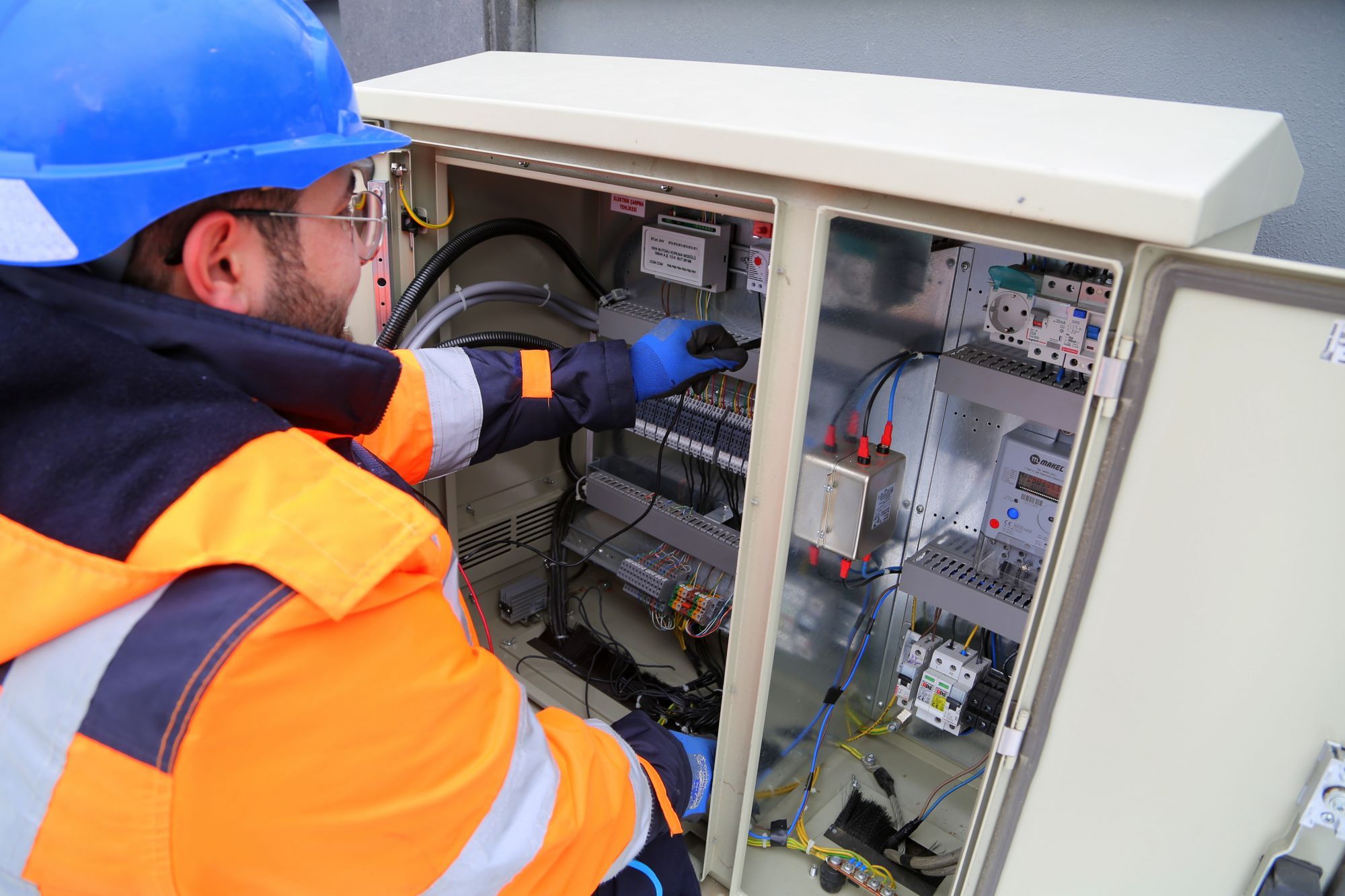 Técnico com capacete azul e casaco de alta visibilidade a trabalhar num quadro eléctrico exterior